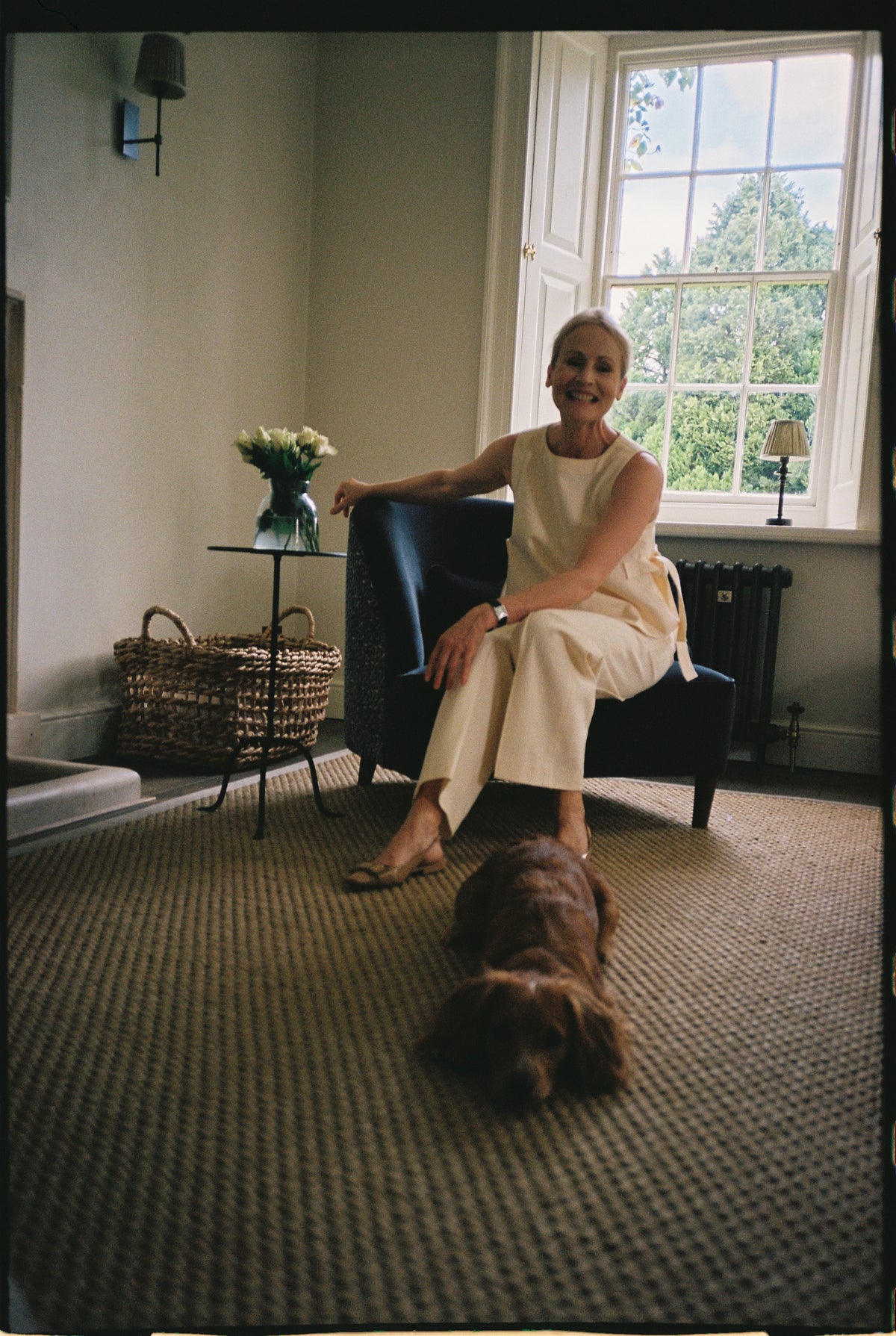 A woman sitting in an upholstered armchair by a sash window, with a dog lying on the rug in front of her.