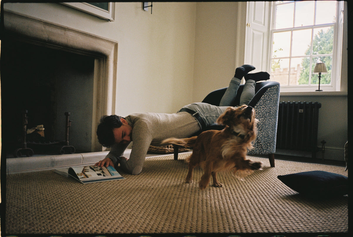 A man lying on the floor of a sitting room playing with a dog, beside an upholstered armchair.