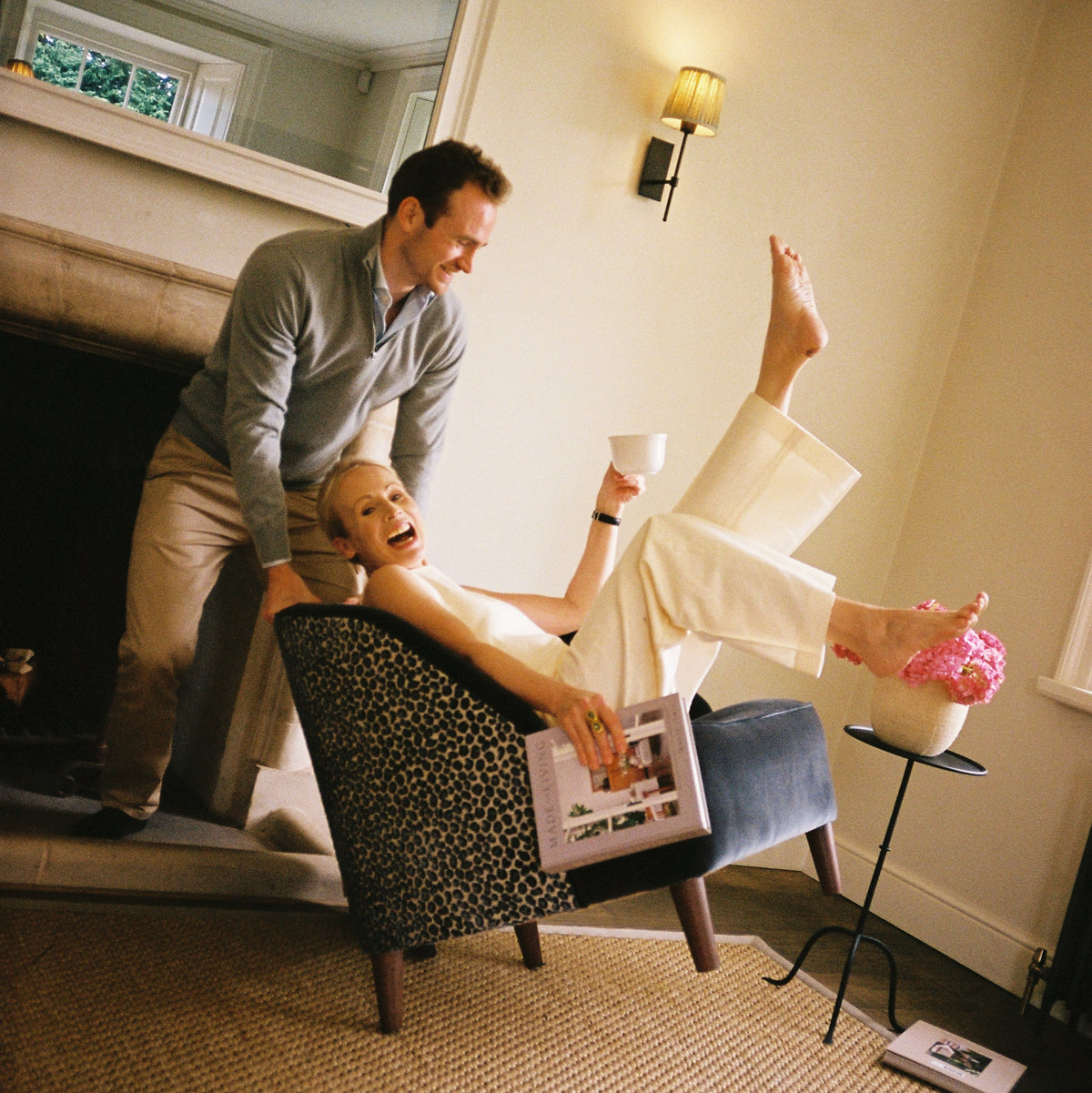 A woman laughing as she is tipped back in an armchair upholstered in a spotted print, in a panelled sitting room.