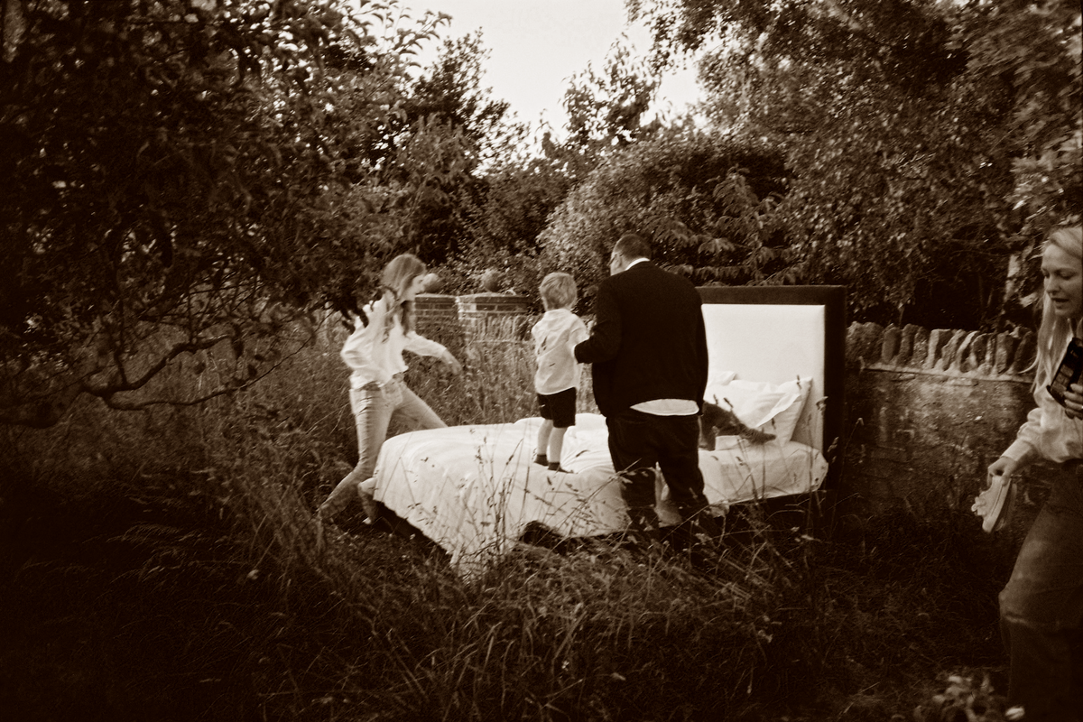A family in a walled garden, with a Muse & Maker upholstered headboard and bedding set out in the long grass.