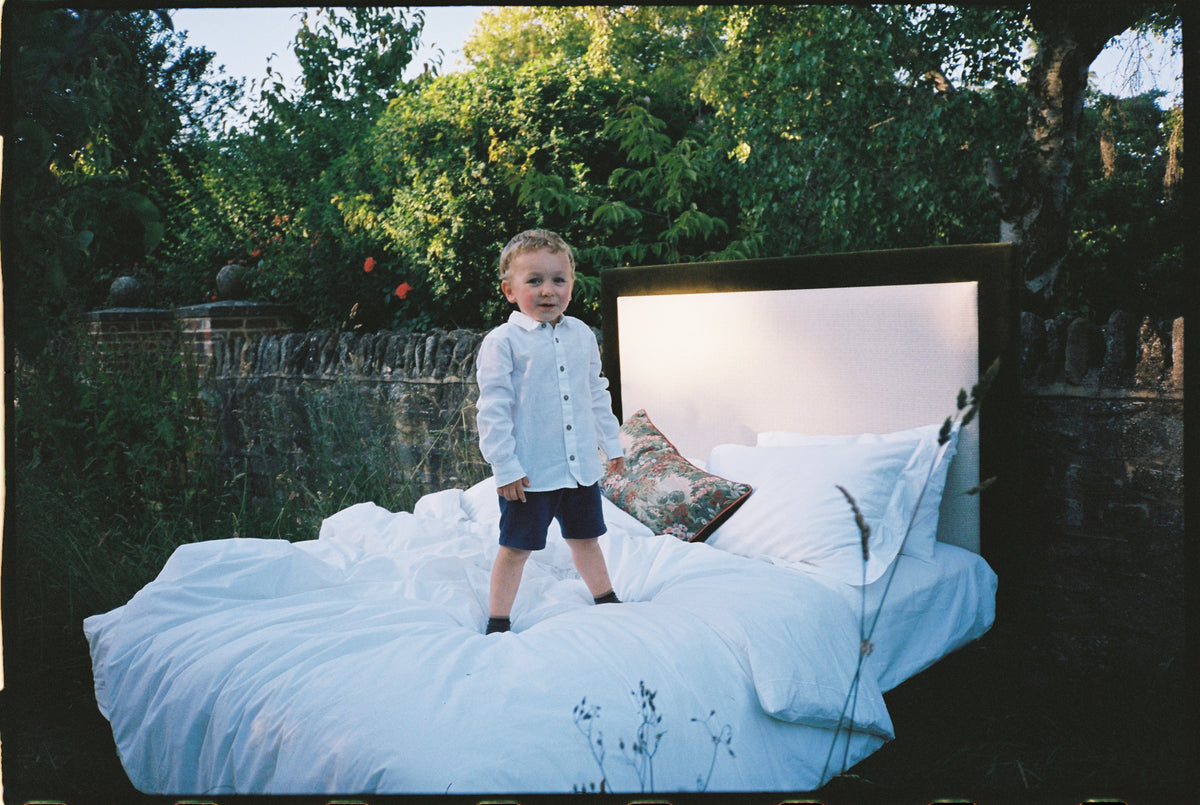 A small boy standing on a bed made up in white linen, set out in a garden.