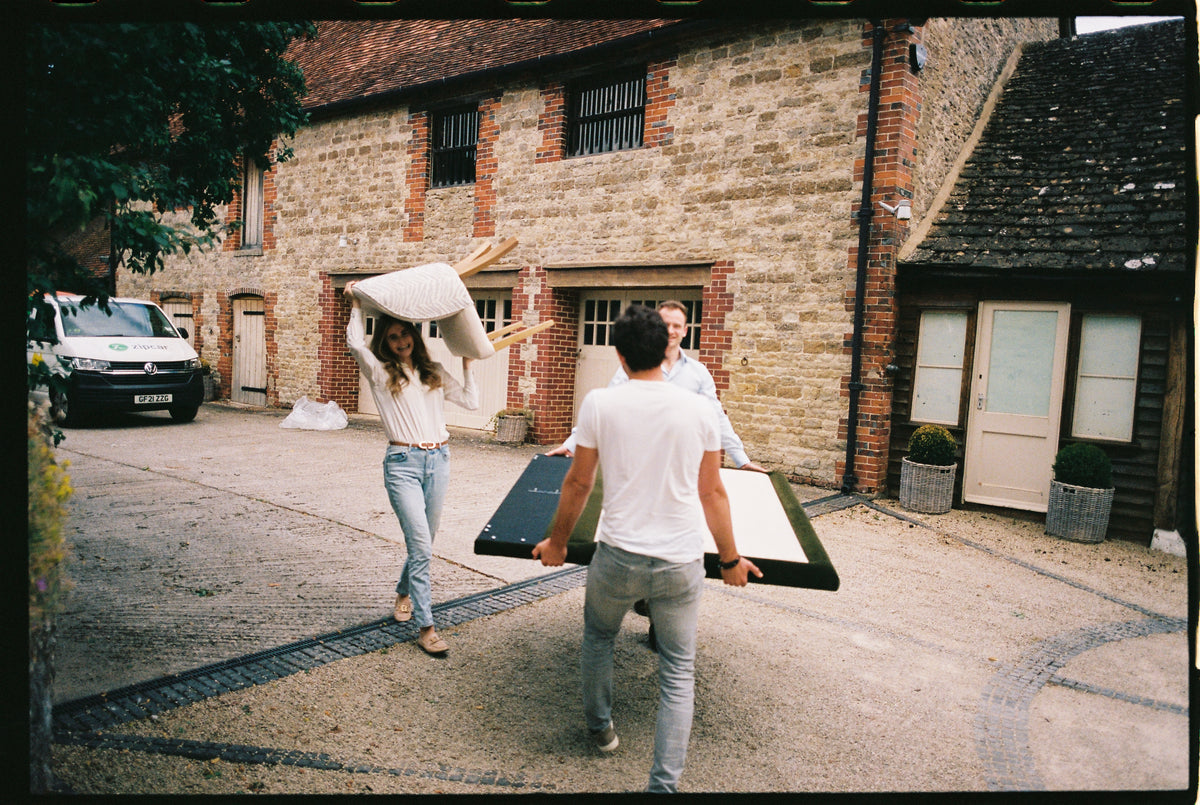 Clara Forstner carrying an upholstered chair across a gravel drive while two men carry a headboard towards a stone house.