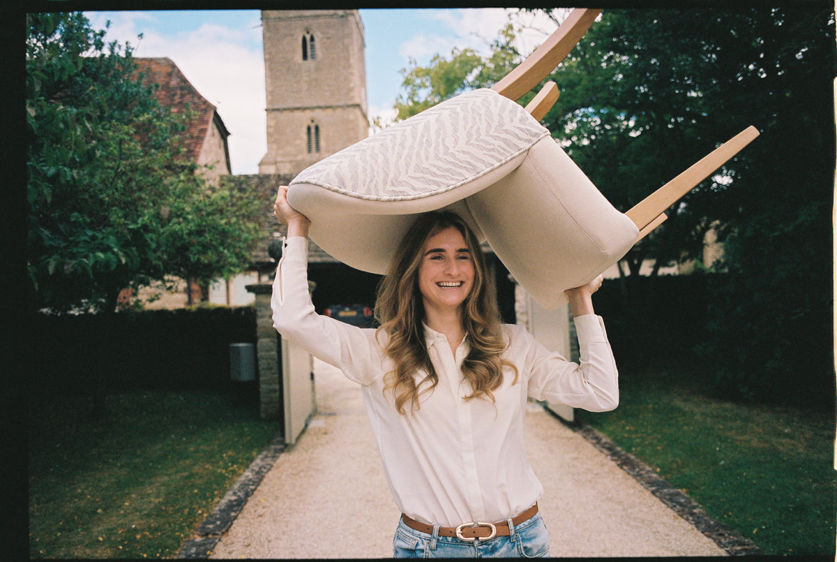 Clara Forstner, founder of Muse & Maker, laughing as she carries an upholstered chair on her head up a garden path.