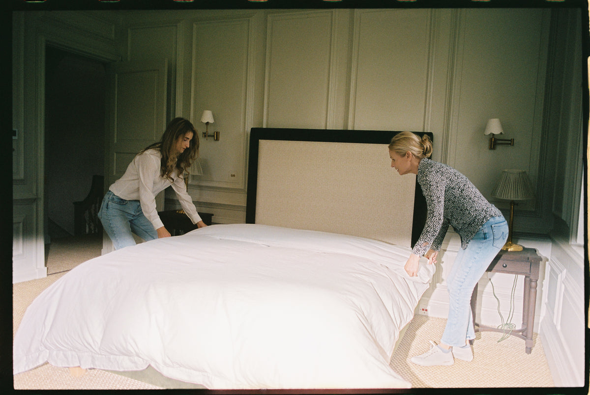 Two women making up a bed with white linen in a panelled bedroom.