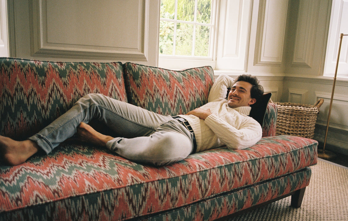 A man resting on a Muse & Maker sofa upholstered in a coral and green flamestitch fabric, in a panelled room.